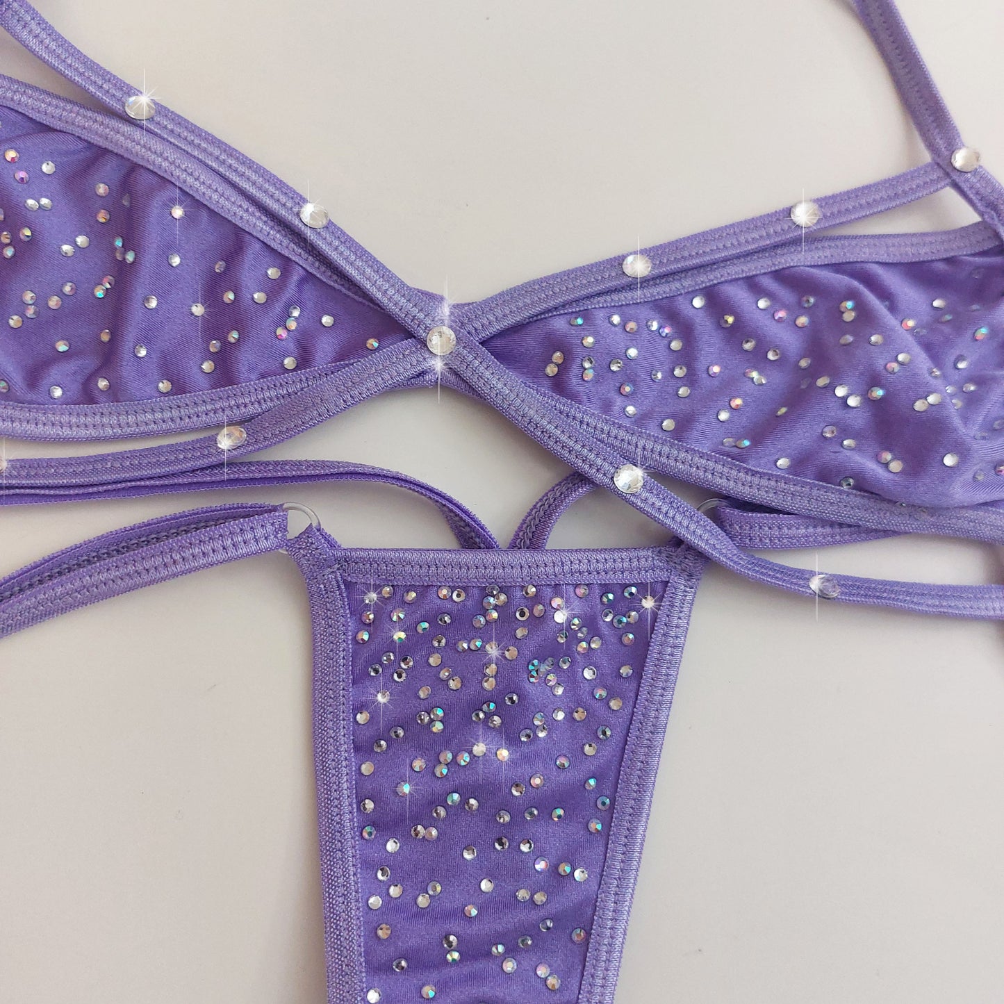 Purple strappy bikini top and thong set embellished with rhinestones, laid flat on a light beige background.